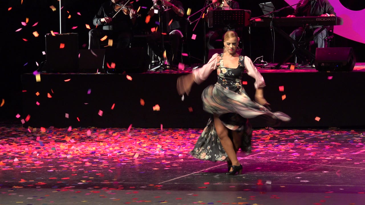 Female dancer in a floral dress spins near center stage in Cabo San Lucas, Mexico, with falling confetti, magenta lighting, and musicians partly visible behind her.