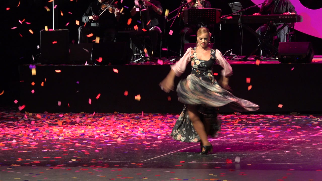 Female dancer continues her stage routine in Cabo San Lucas, Mexico, with her skirt blurred by motion, confetti scattered across the floor, and band equipment in the background.