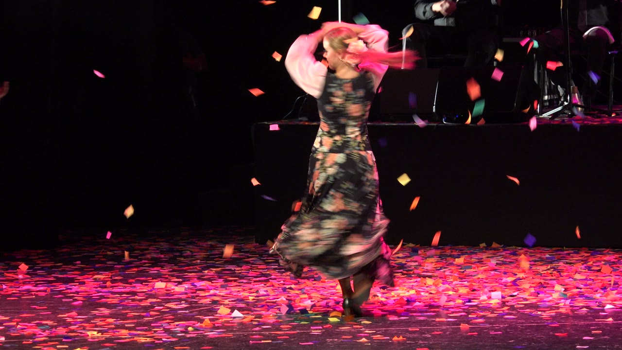 The dancer is seen from behind on the Cabo San Lucas stage with both arms lifted, her floral dress blurred by movement and colored confetti crossing the dark background.