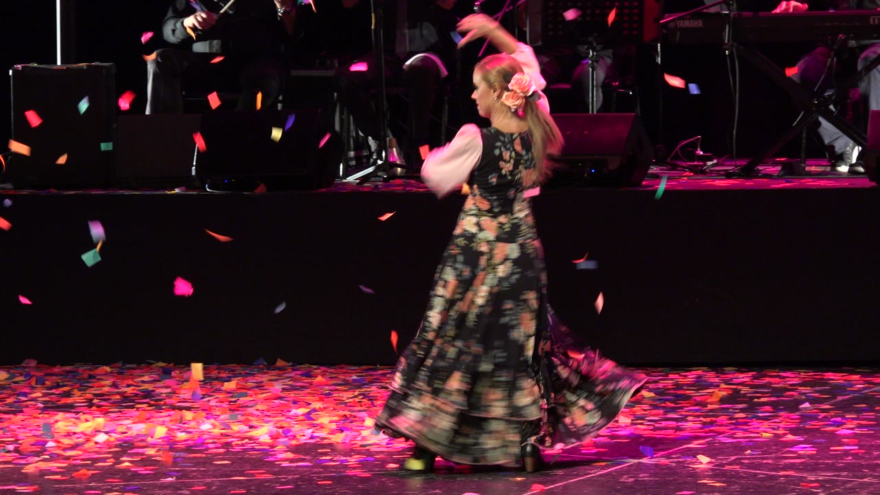 In Cabo San Lucas, Mexico, the performer is seen from behind and slightly to the side as loose confetti hangs in the air above the dark stage floor.