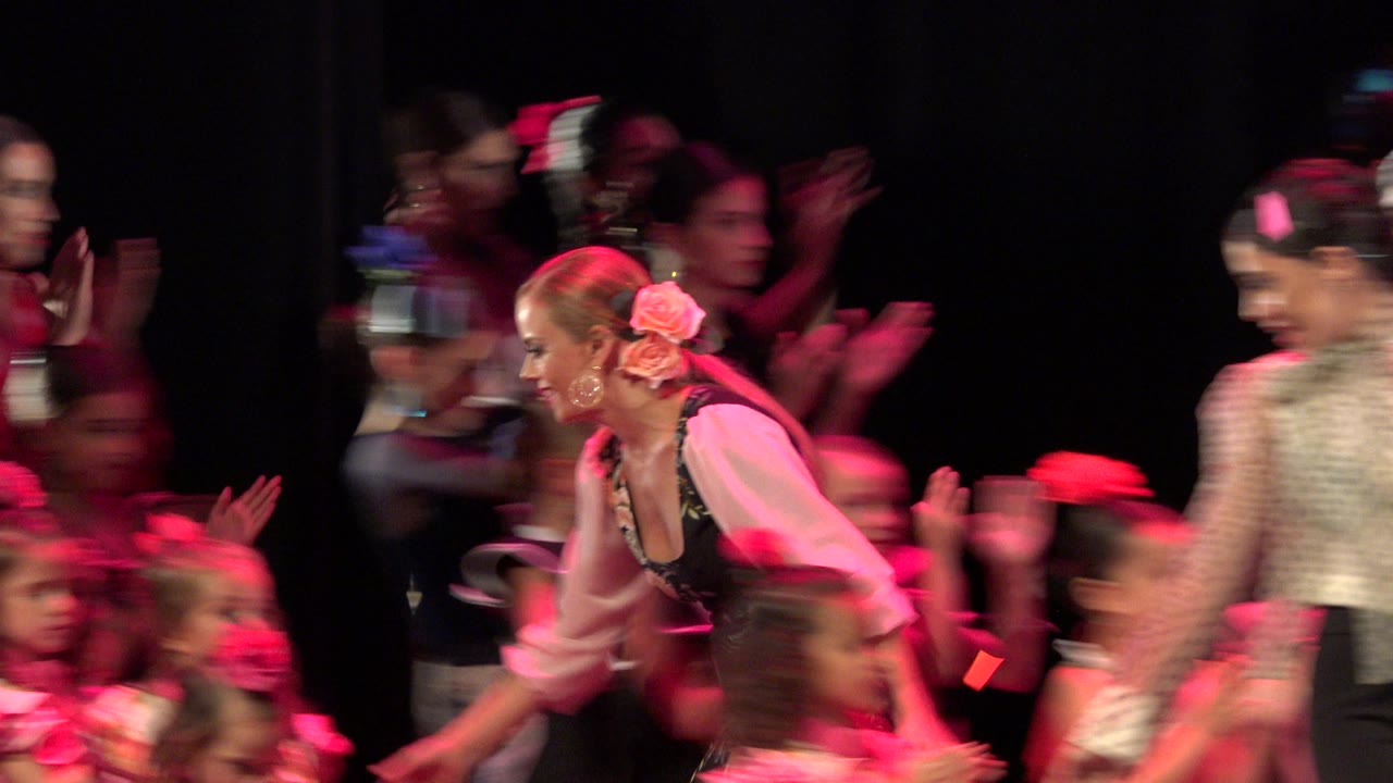An adult dancer in a floral outfit bends toward children onstage in Cabo San Lucas as other performers clap in the red-lit background.