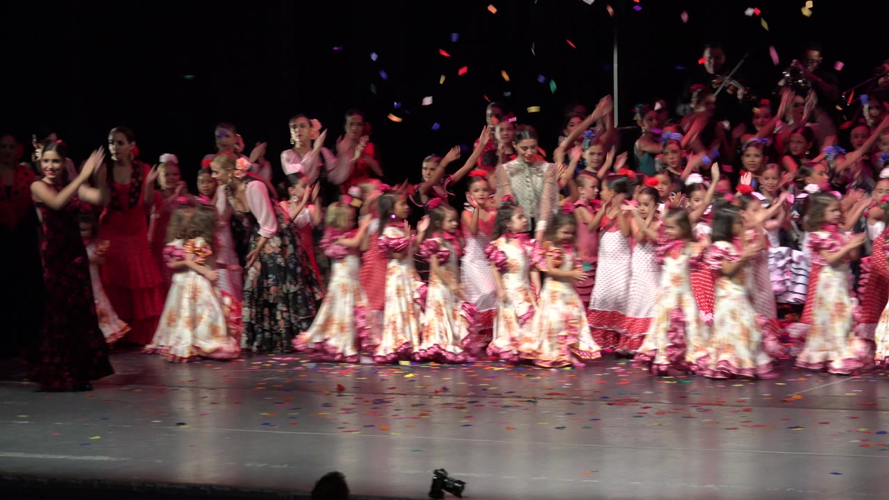 Wide stage view of children and adult performers clapping under falling confetti.