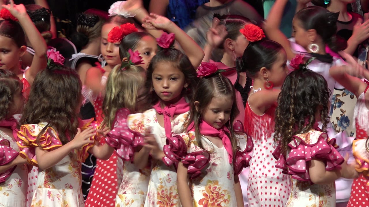 Children in Cabo San Lucas, Mexico perform with arms raised and faces turned in different directions as pink stage light falls across floral dresses and red hair flowers.