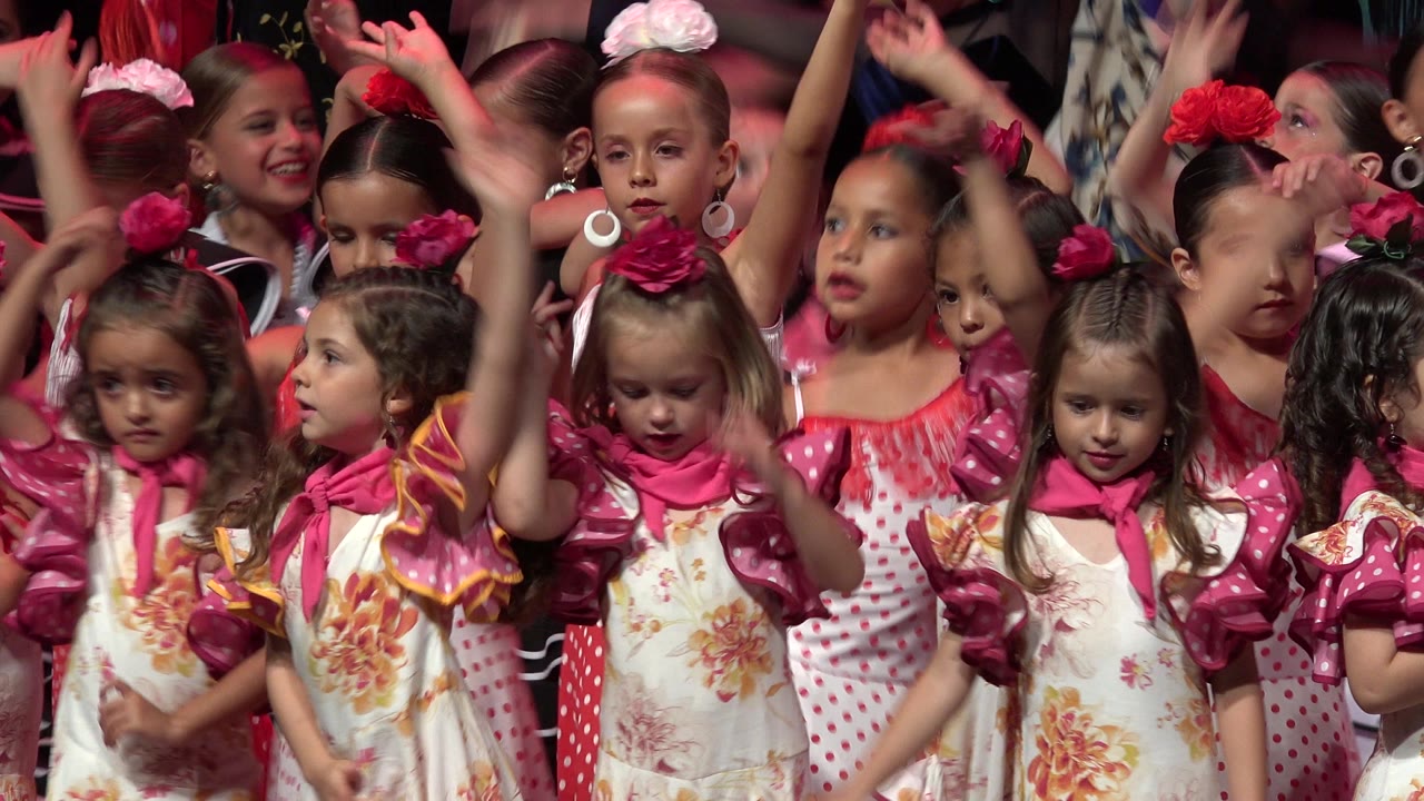 Young girls in Cabo San Lucas stand shoulder to shoulder in pink and floral dance costumes as several children raise their arms at the start of the clip.