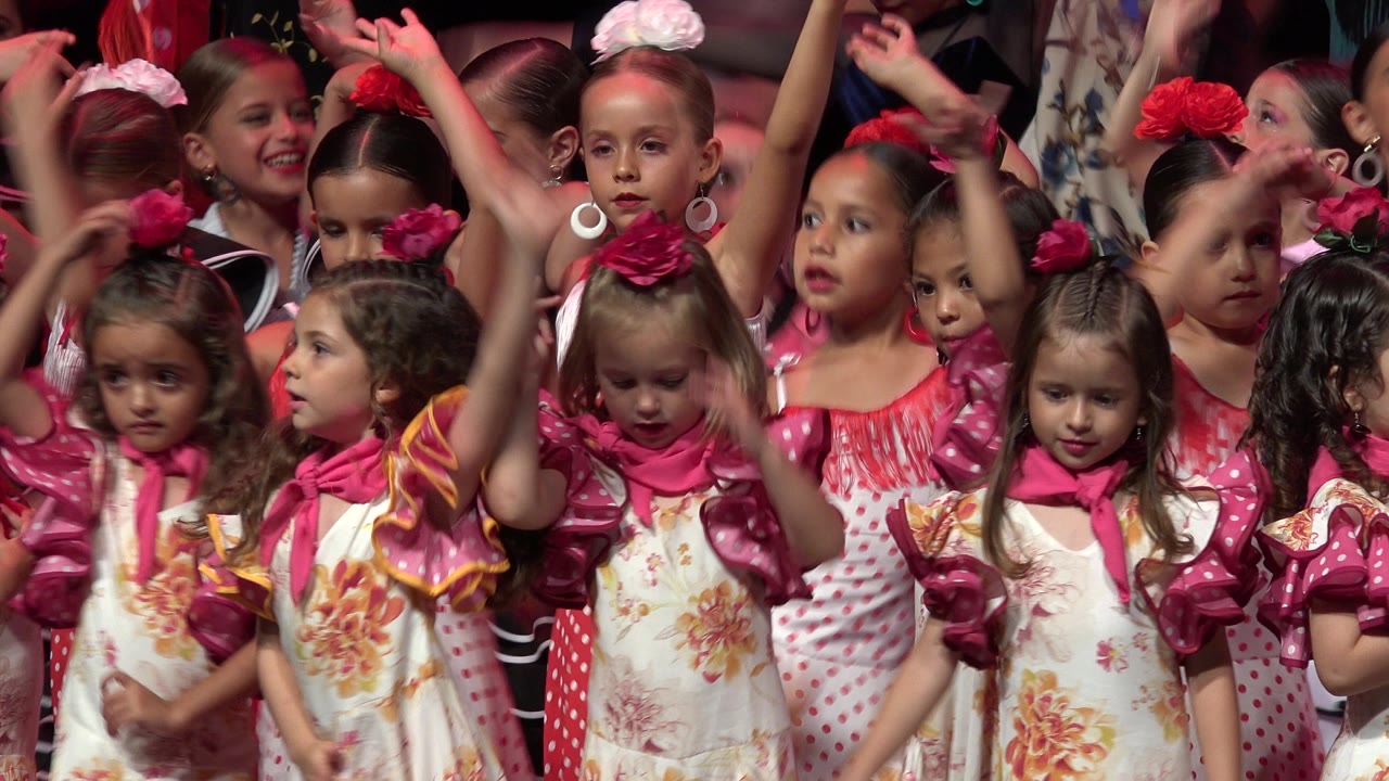 Girls in red polka dot and pink ruffled costumes perform in Cabo San Lucas, with some looking sideways while others lift their hands above the group.