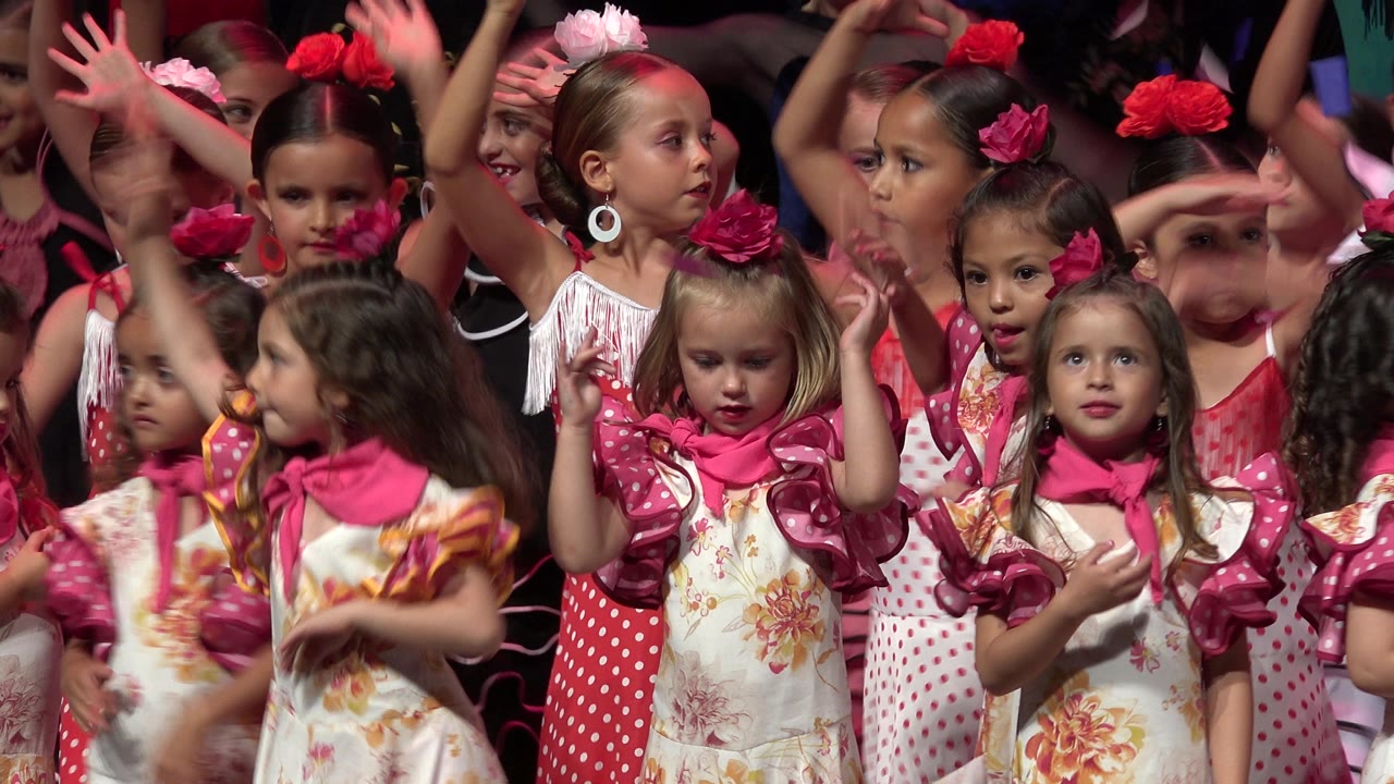 Girls in coordinated pink costumes crowd the stage with hands moving overhead.