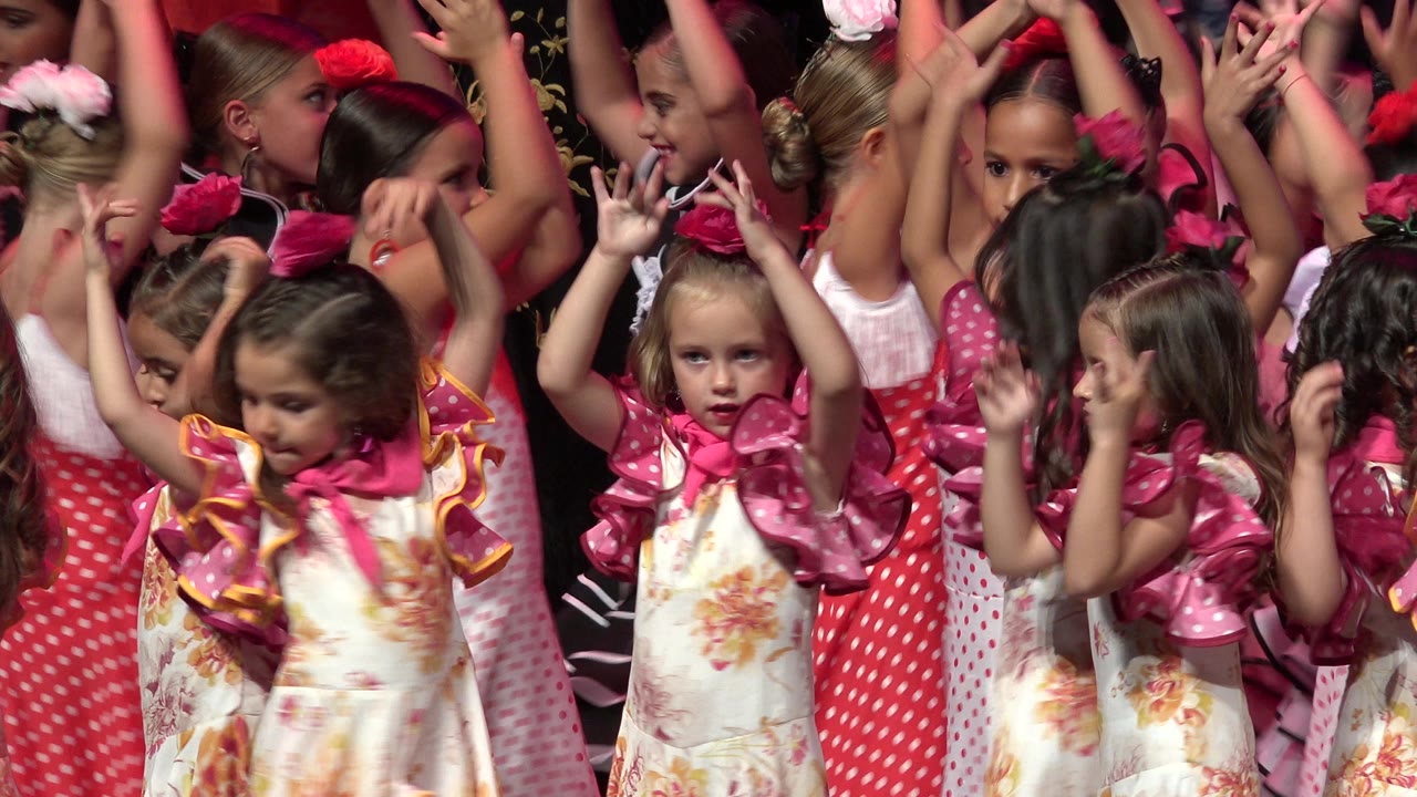 At 0:00 in Cabo San Lucas, young girls in pink and white dresses stand close together on an indoor stage with arms lifted under red stage light.