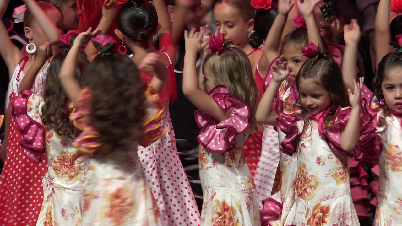Girls in floral dresses move their raised arms on a stage.