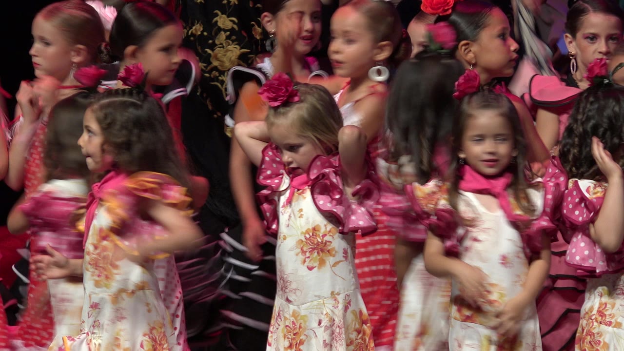 Several young girls in Cabo San Lucas face different directions while singing or speaking, with pink dotted costume sleeves and floral dresses filling the frame.