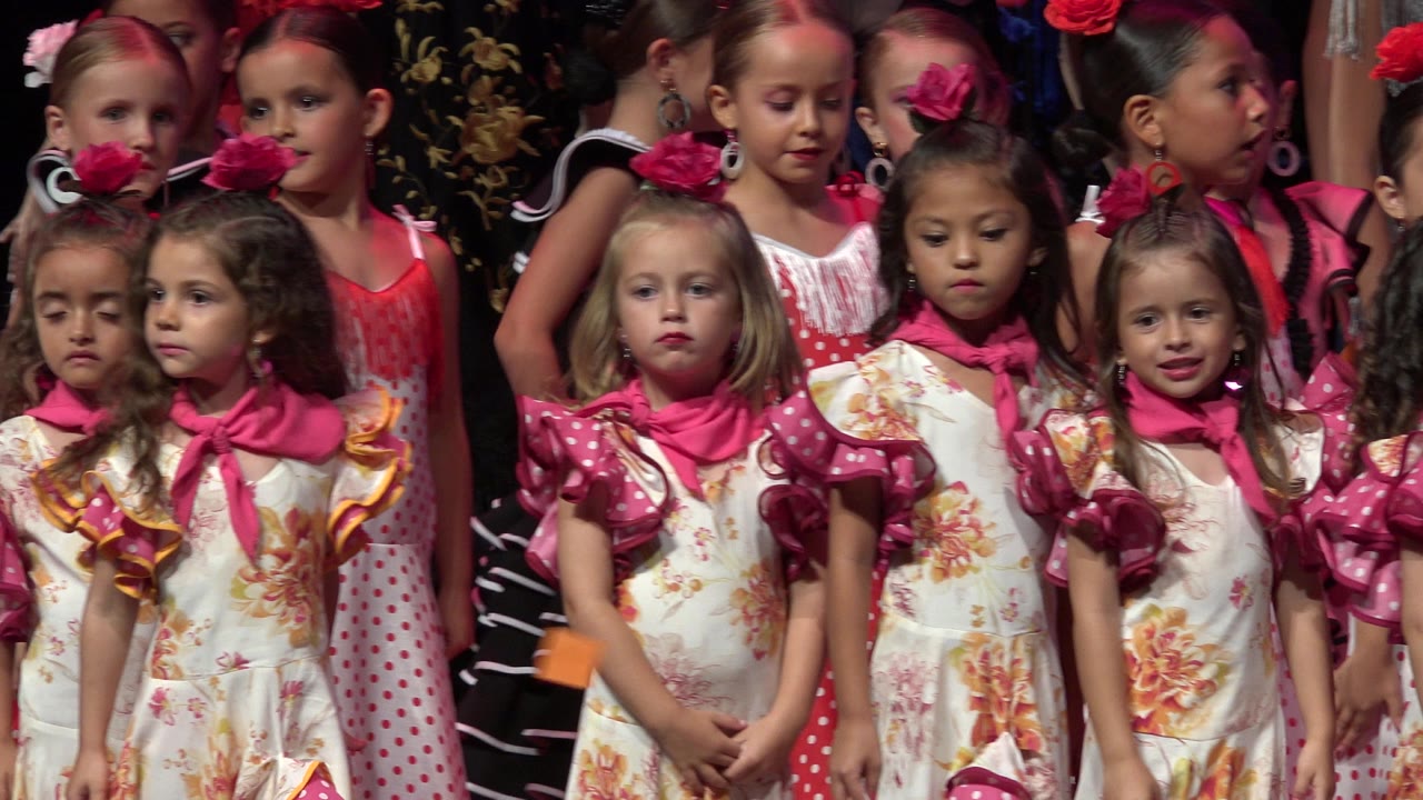 Young girls in floral dresses and pink scarves stand in front of other costumed children on a lit stage in Cabo San Lucas, Mexico.