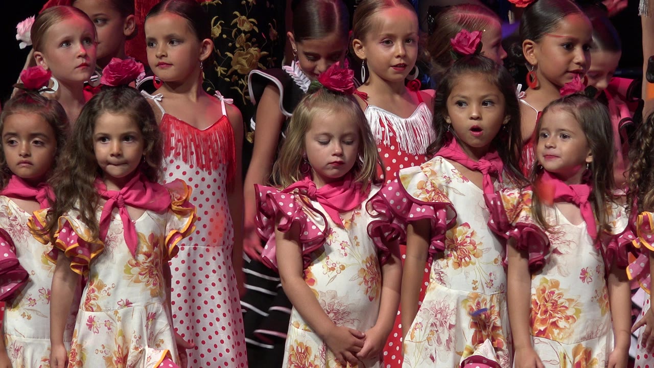 Young performers face the camera in a tight stage grouping, with pink scarves, floral fabric, and hair flowers prominent in the frame.