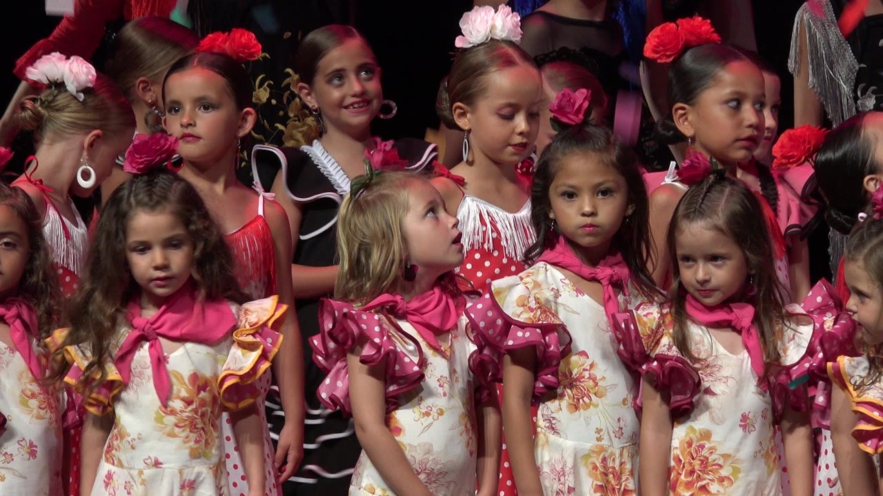 Young girls in colorful ruffled costumes stand closely together in Cabo San Lucas, Mexico, with visible faces and a dark performance background.