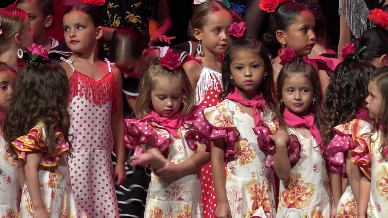 Young girls in pink bows and ruffled performance outfits stand close together during a Cabo San Lucas, Mexico stage moment, with adult figures partly visible behind them.