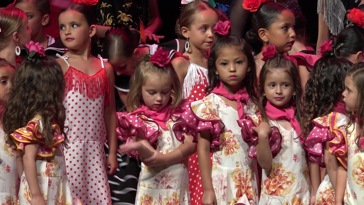 Several young girls in pink and white costumes face different directions on a crowded Cabo San Lucas, Mexico stage, with red-pink light across their hair and faces.