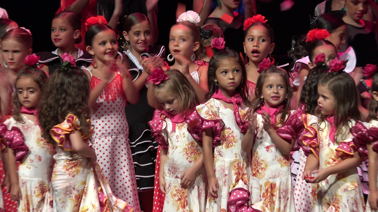 Young children in floral dresses and pink hair accessories clap on a stage in Cabo San Lucas, Mexico, with older performers partly visible behind them.