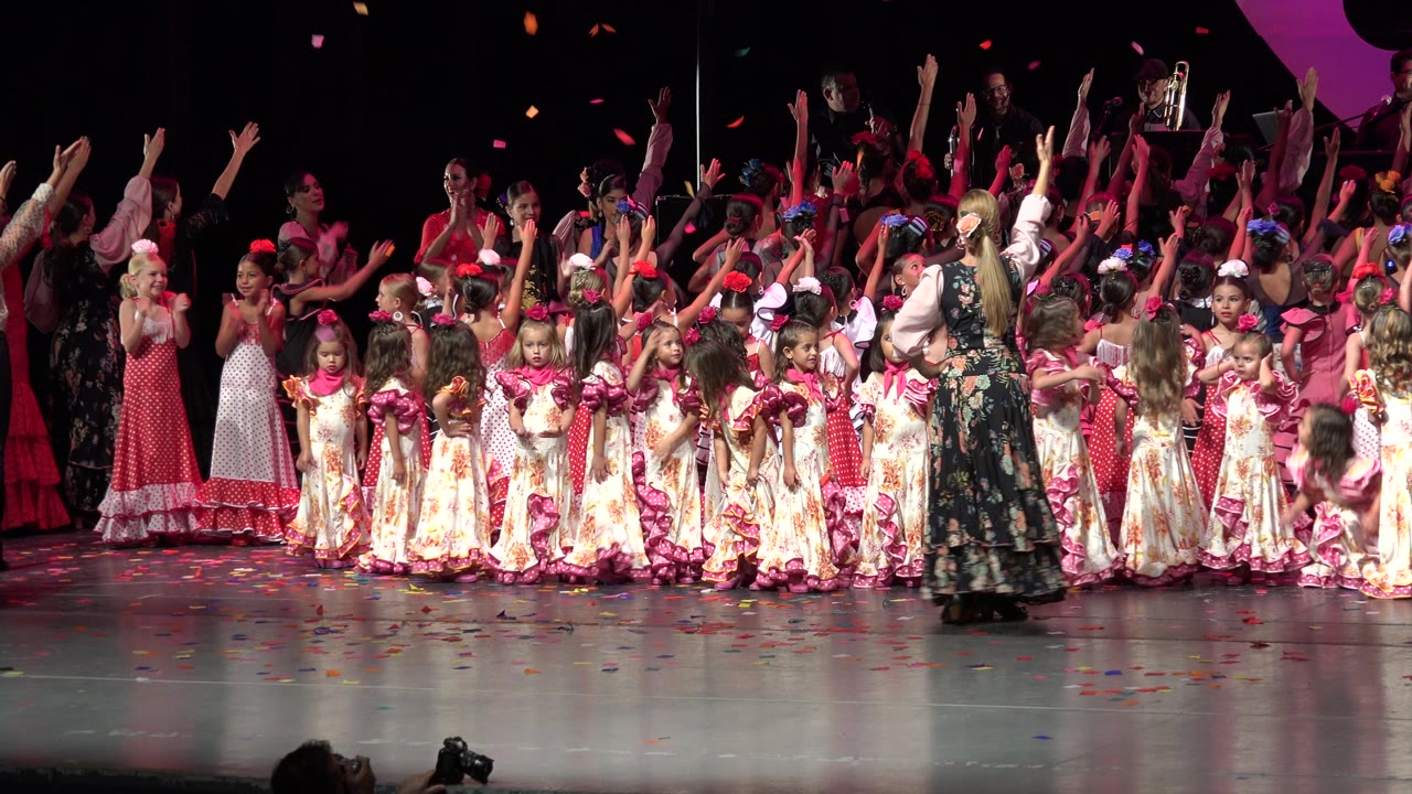 Girls in colorful dresses stand on a stage with raised hands.