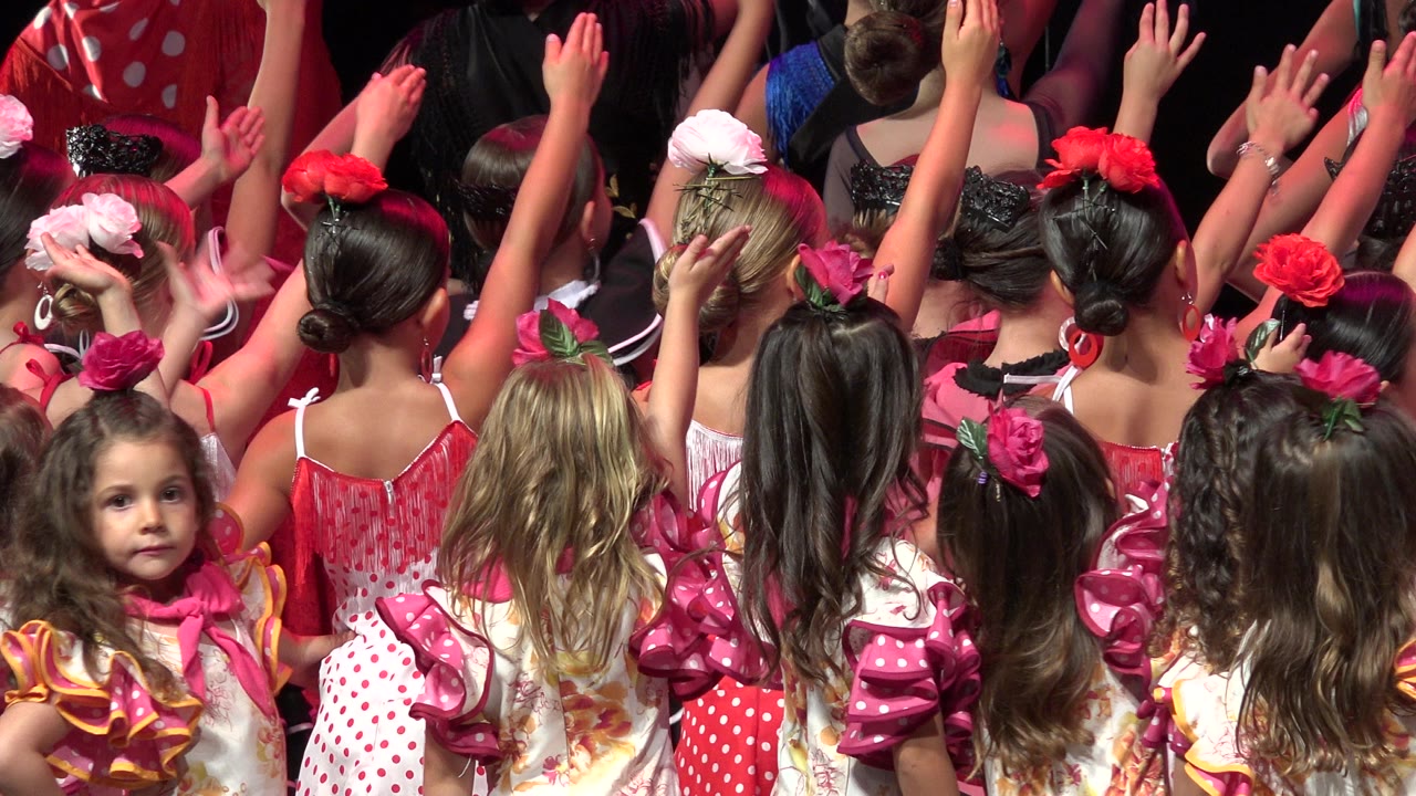 At 0:01, the performers remain tightly grouped with raised arms, floral hairpieces, and bright pink costume details under warm stage light in Cabo San Lucas, Mexico.