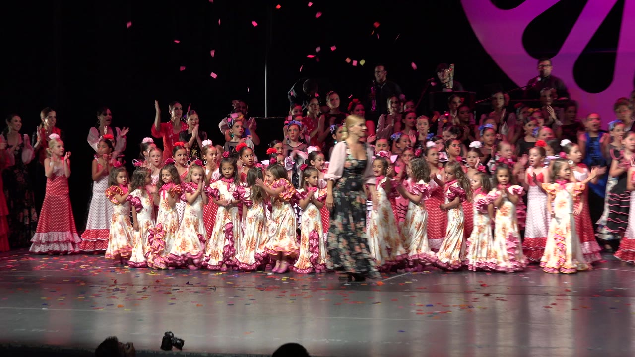 Wide stage with children and adults applauding around a woman in red and gold costume.