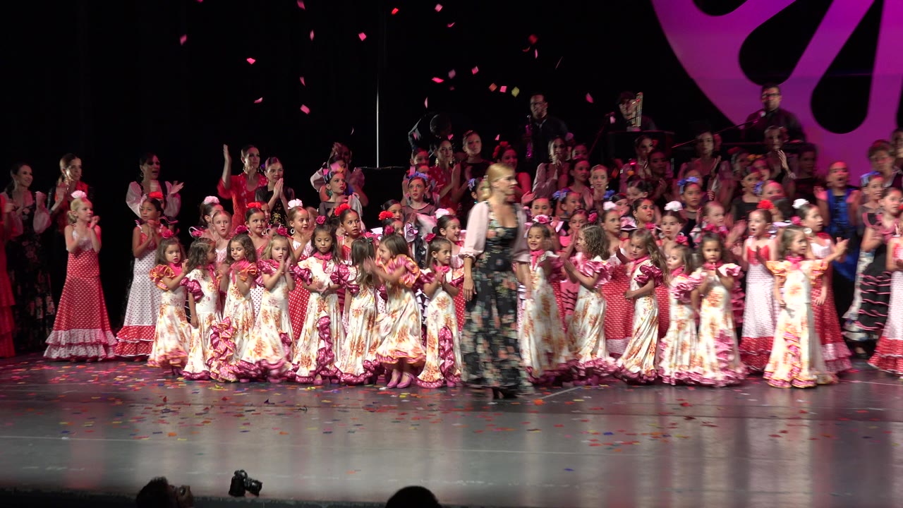 In Cabo San Lucas, Mexico, performers fill the stage as children in pale floral costumes stand in front and adults in red, black, and patterned dresses applaud behind them.