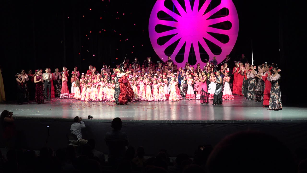Wide stage full of performers under a magenta fan-shaped backdrop.