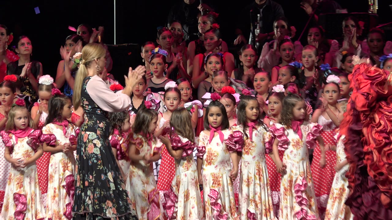 Young girls in colorful dresses stand on a stage as an adult woman applauds beside them.
