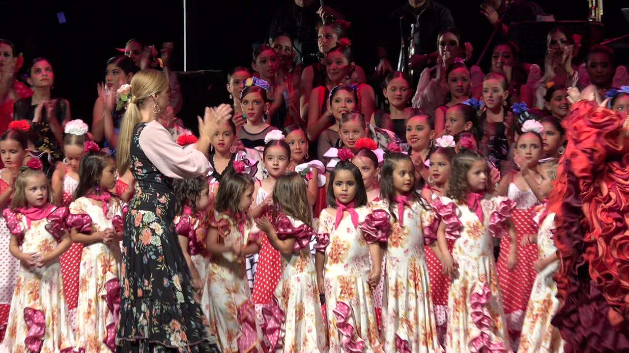 In Cabo San Lucas, Mexico, the group of children remains clustered under pink stage light while the adult woman stands near the front row and spectators fill the background.