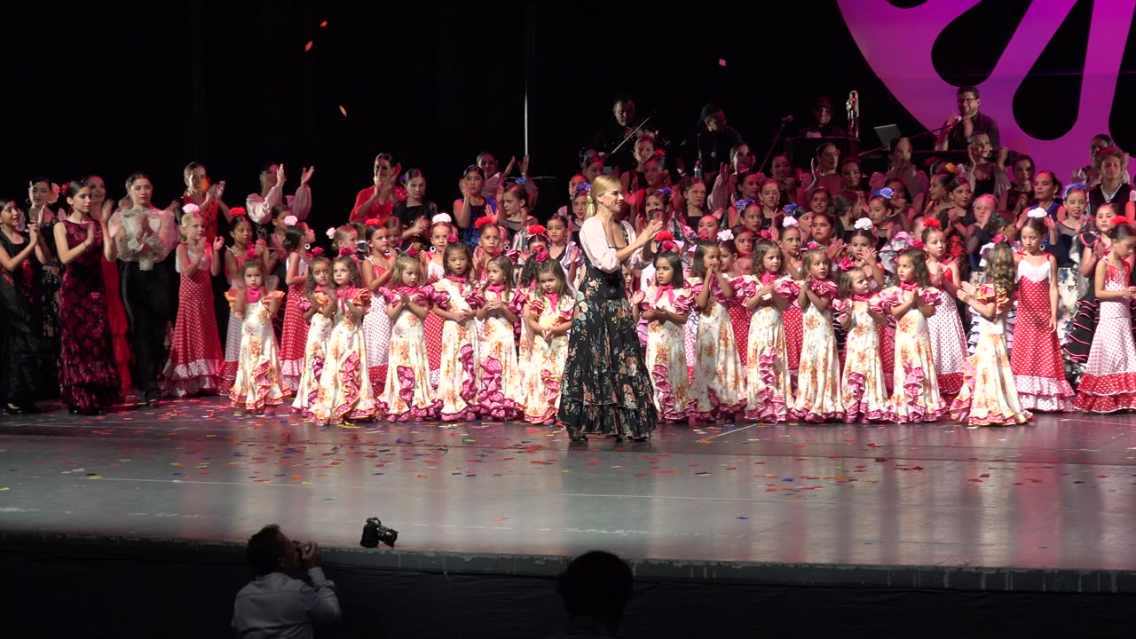 Wide view in Cabo San Lucas, Mexico of children in floral and red dresses lined across a stage while adults and older performers clap in the background.