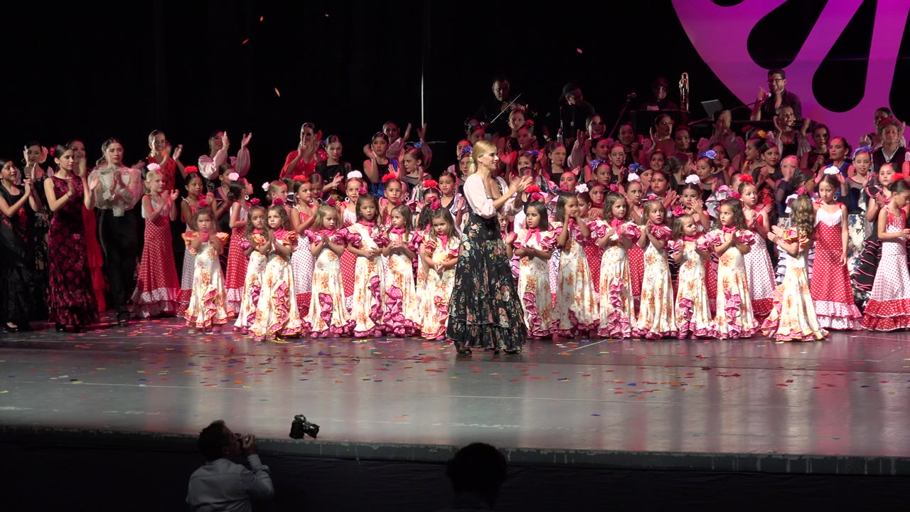 Wide Cabo San Lucas stage frame with a blonde woman clapping near the front row of children as the larger group stands behind under red-pink light.