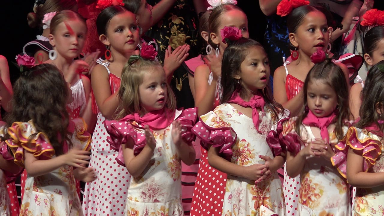 Girls in pink and white dresses clap on an indoor stage.