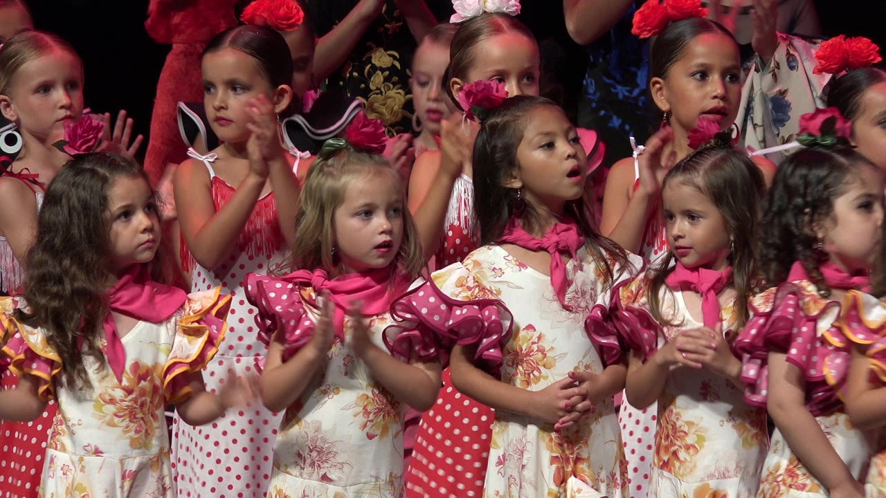 Children in red and floral dance costumes stand on a lit stage.