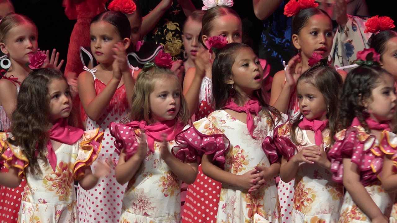 A tight stage view in Cabo San Lucas shows young girls in floral dresses, red polka dots, and flower headpieces looking in different directions.