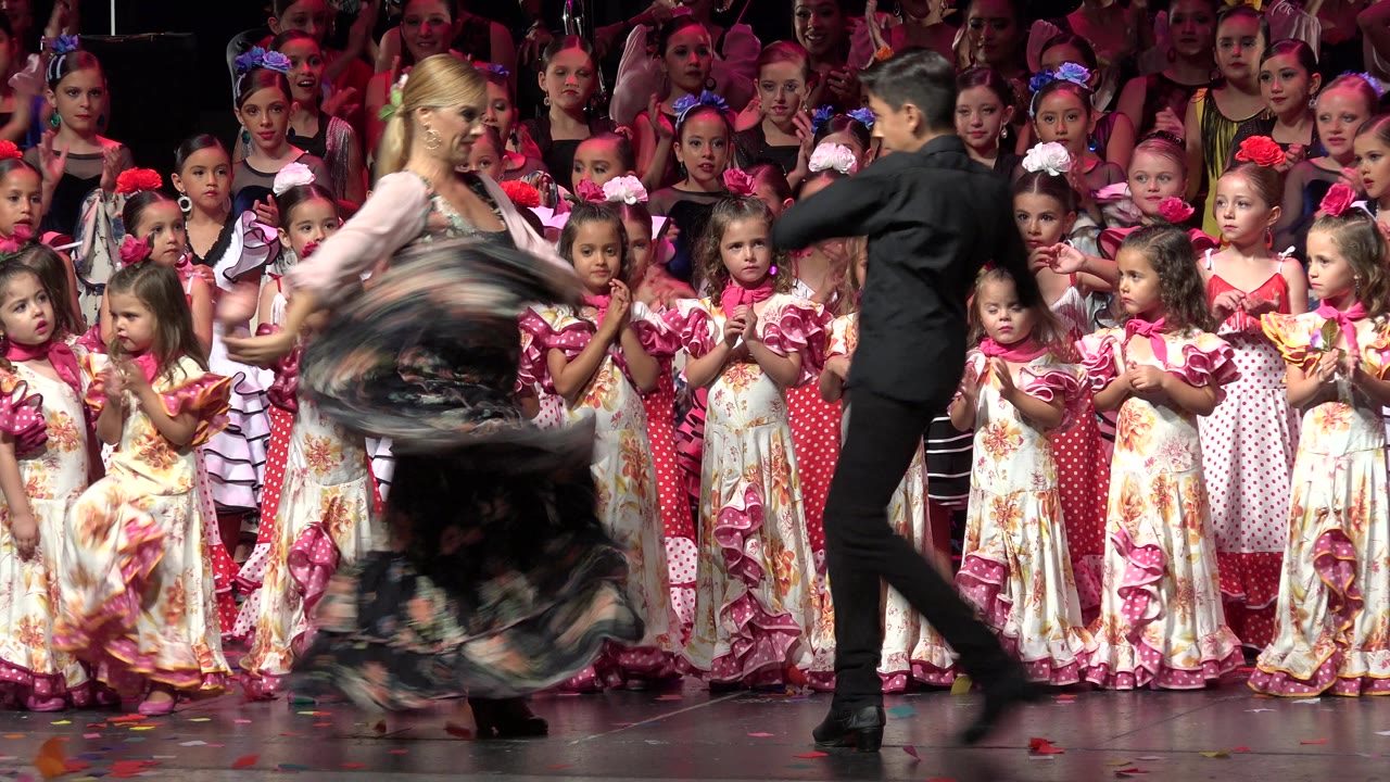 In Cabo San Lucas, Mexico, the woman spins her dark floral skirt while the man in black steps beside her and children in coordinated stage costumes watch from behind.