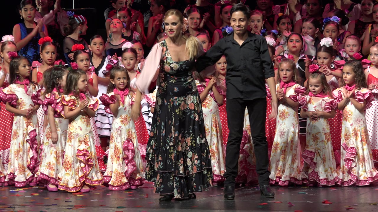Two adult presenters stand before children in pink and cream stage costumes.