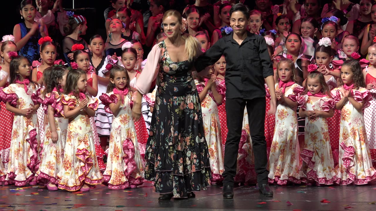 The Cabo San Lucas stage scene holds on the woman and man standing together as children in ruffled pink and cream outfits look forward behind them.