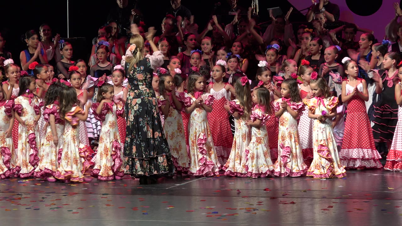 Young dancers in floral and red costumes clap on a lit stage in Cabo San Lucas, Mexico, with an adult in a dark floral dress facing the group.