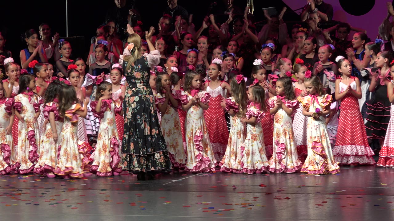 A group of children in coordinated dresses clap beside an adult on a bright stage in Cabo San Lucas, Mexico, with more performers visible behind them.