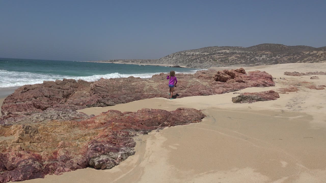 Child in a purple shirt standing on a sandy beach near red rocks and surf.