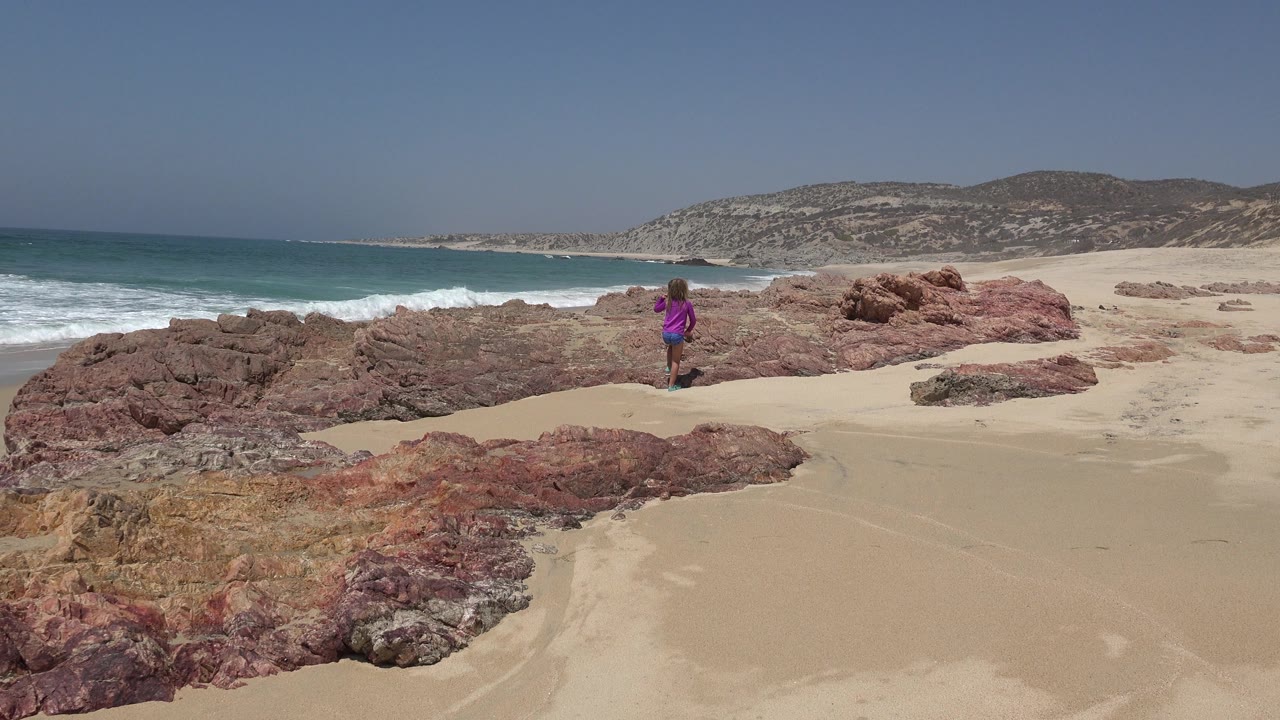 The child remains near the center of the Cabo San Lucas beach frame, standing beside reddish rock ledges while a wave breaks along the left edge of the shore.