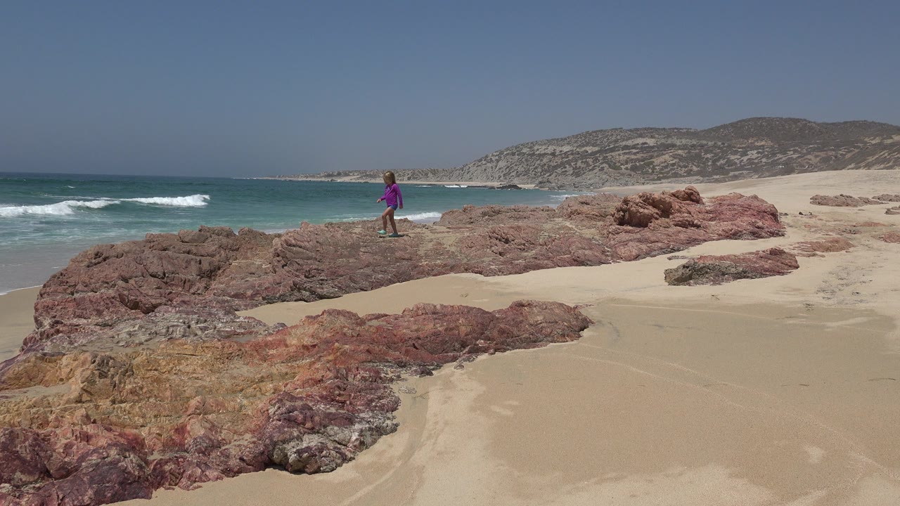 A young girl in a purple top stands on reddish rocks beside surf and open sand in Cabo San Lucas, Mexico.