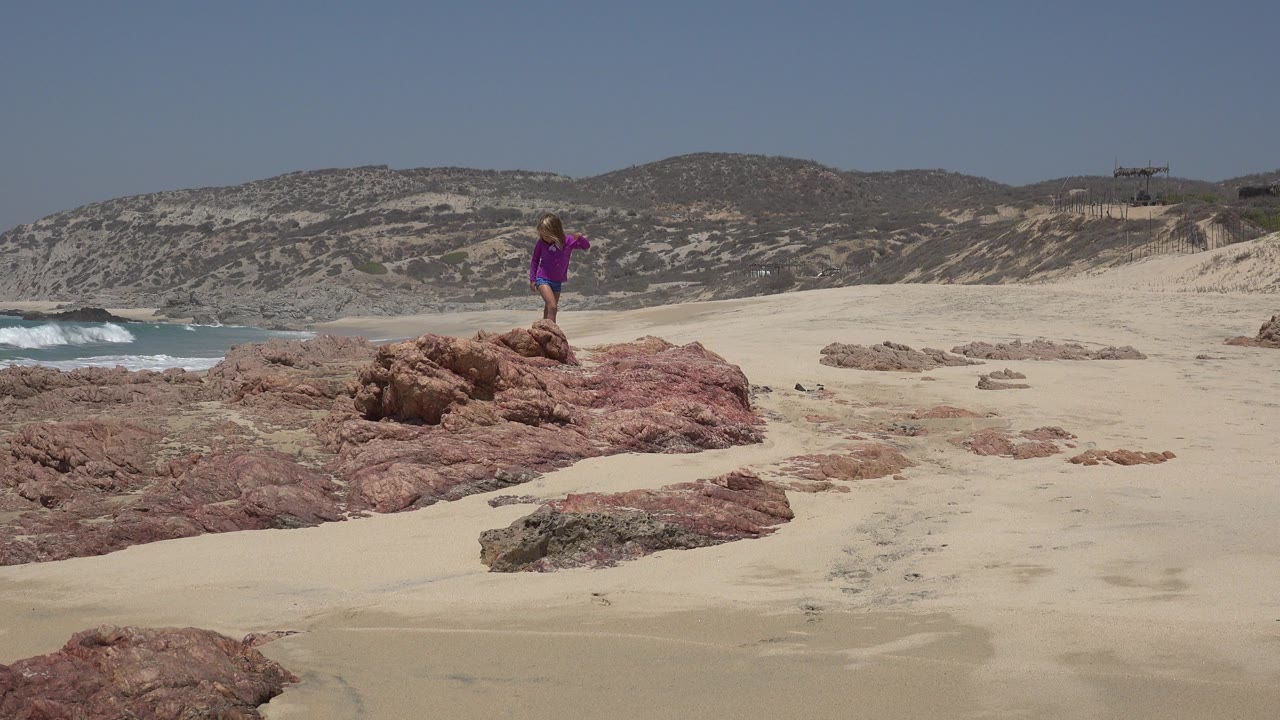Young girl in a pink dress standing on beach rocks near surf.
