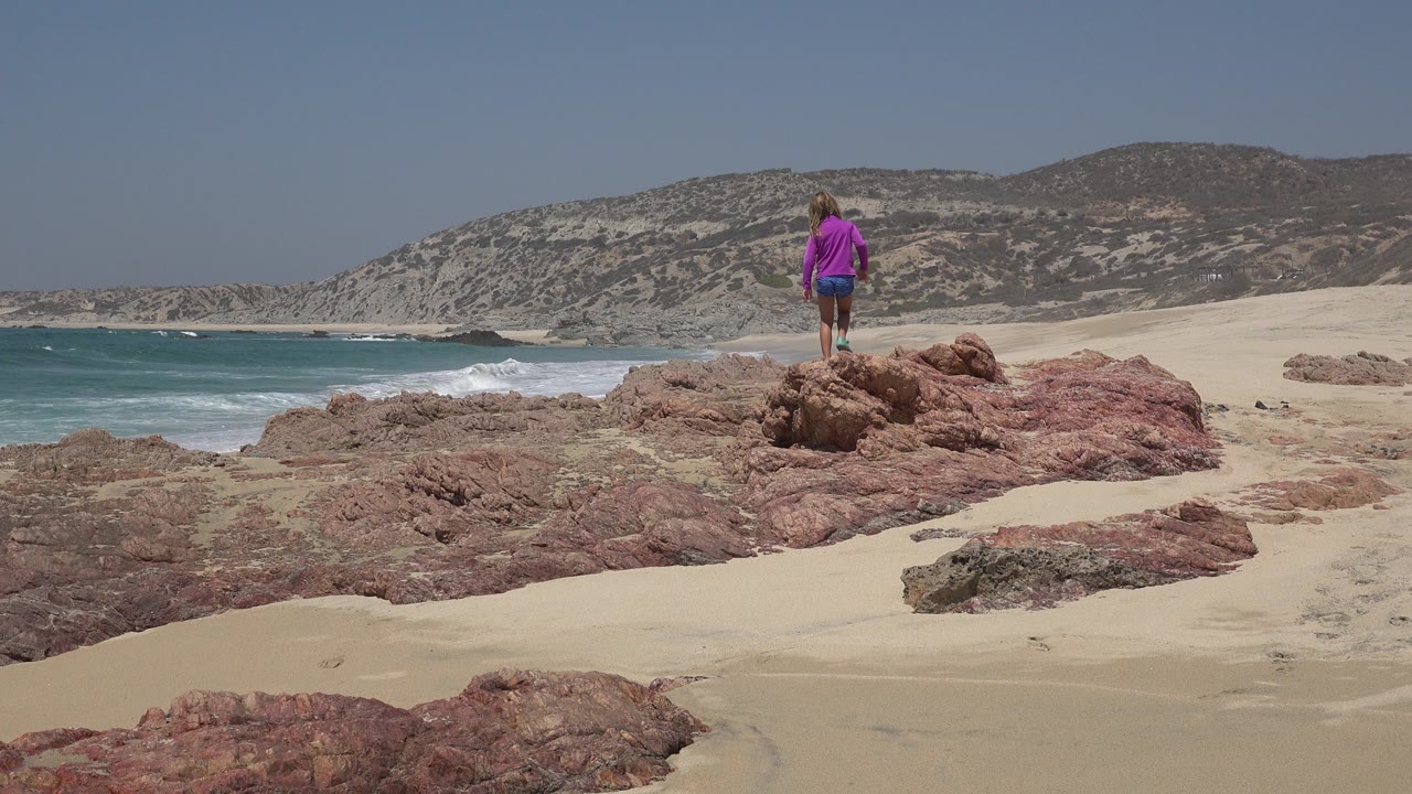 Child standing on reddish beach rocks near ocean surf.