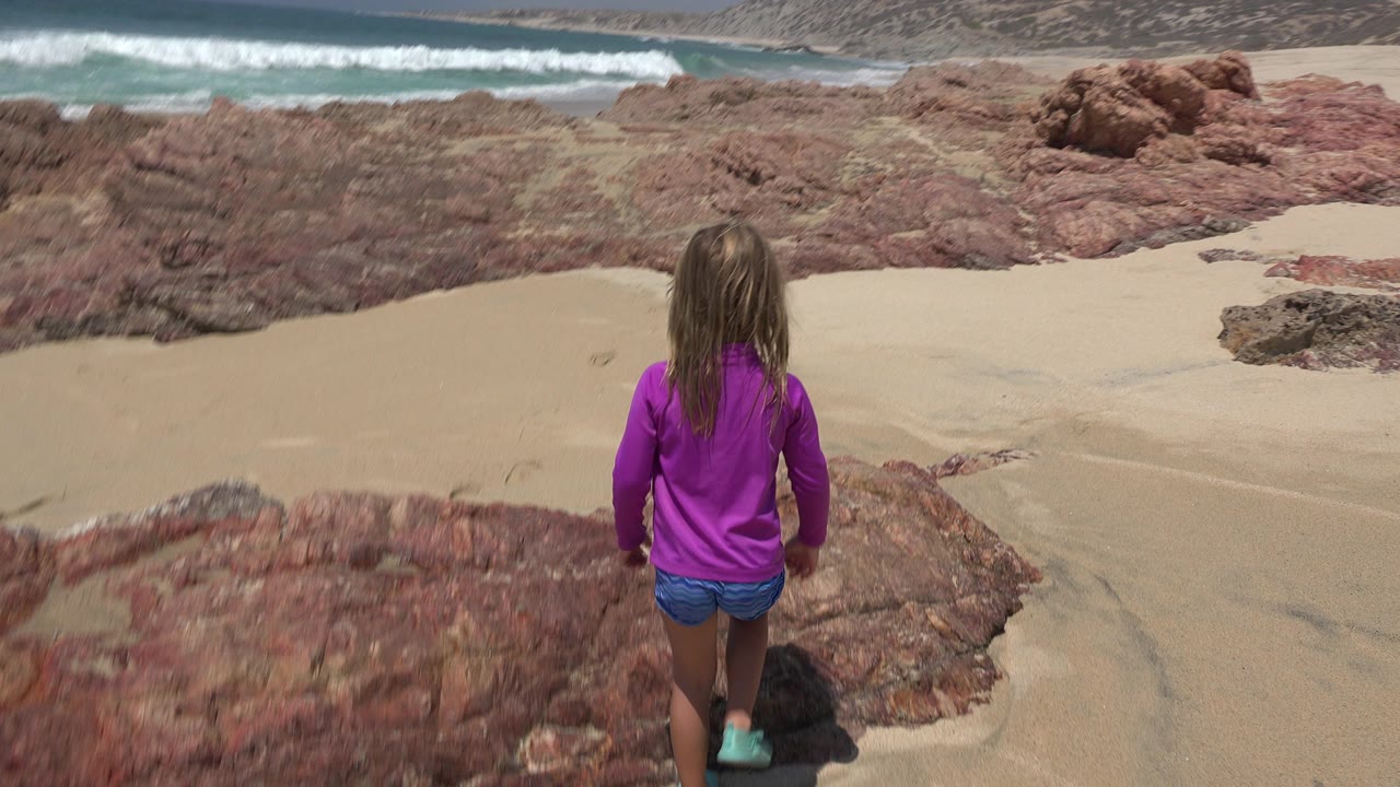 Girl standing on red rocks above a sandy beach and surf.