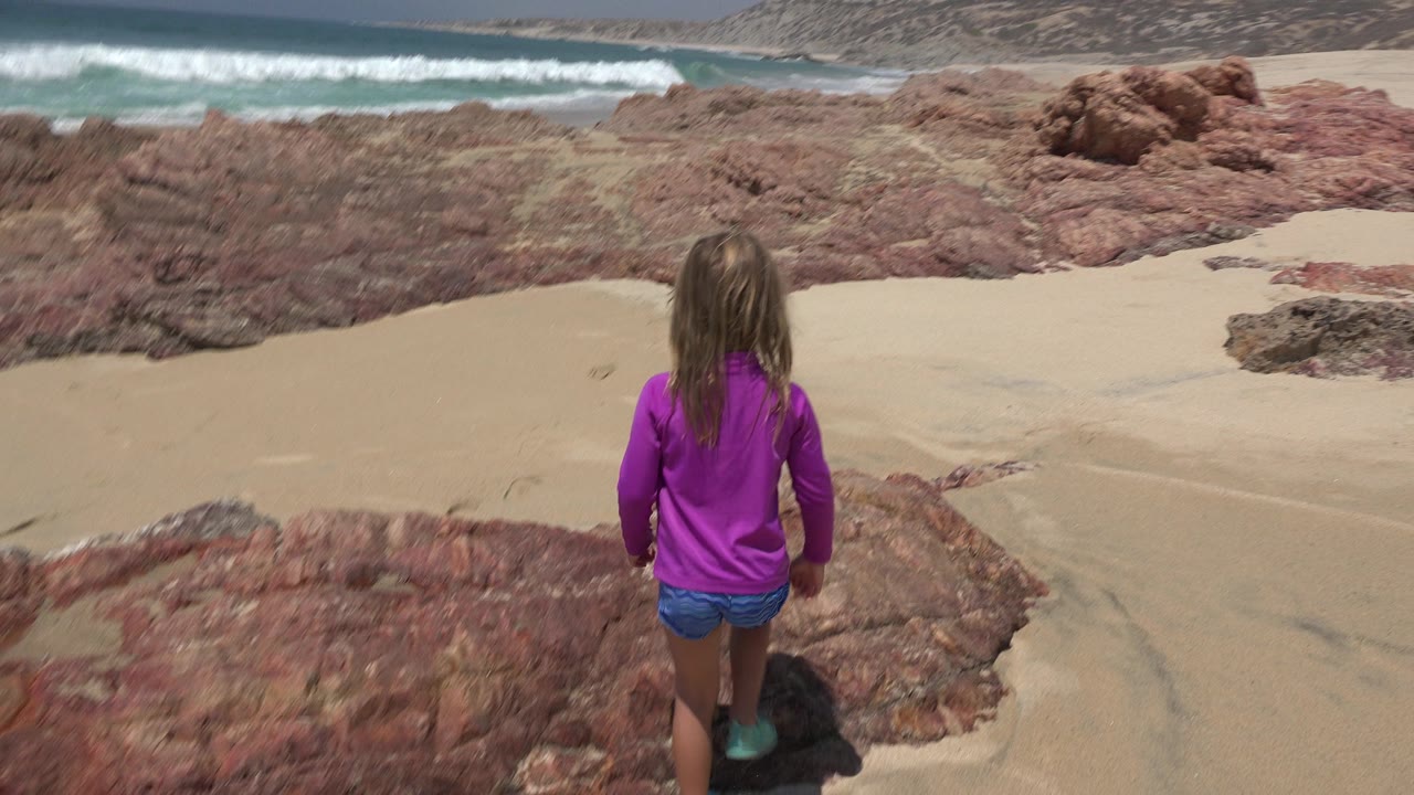 The girl remains centered from behind on the rock ledge as white surf rolls across the beach below in Cabo San Lucas, Mexico.