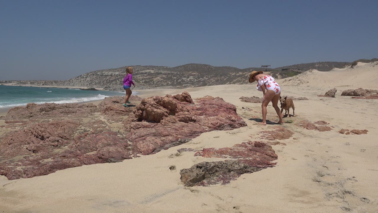 At 0:00 in Cabo San Lucas, Mexico, a child in a purple top stands on beach rocks while a woman in a sun hat bends near the sand, with surf, dunes, and hills behind them.