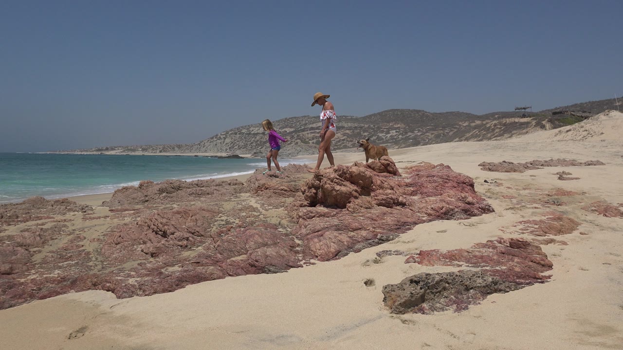 The child steps ahead of the woman on the rocky Cabo San Lucas shoreline, with surf breaking along the left side and low hills across the background.