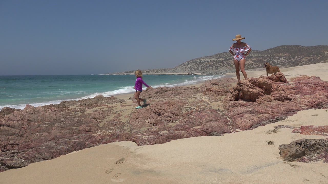 At Cabo San Lucas, the child has moved farther along the rocks toward the center as the woman and small dog remain on the raised reddish stones beside the sandy beach.