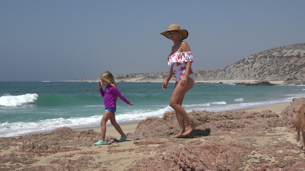 Woman and young girl walking on rocks beside ocean waves.