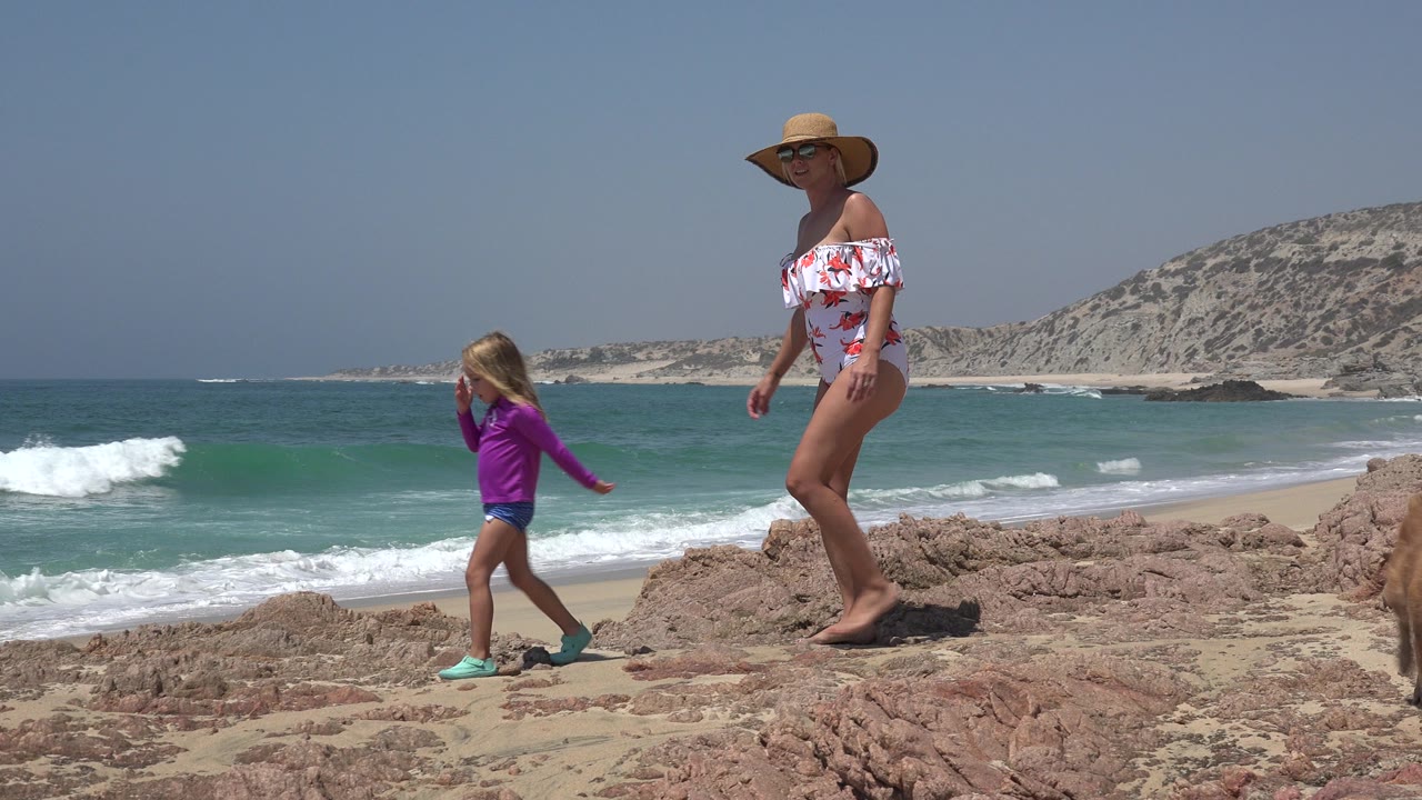 In Cabo San Lucas, Mexico, the girl steps forward on the rocks while the woman walks behind her, with white surf rolling toward the sandy shoreline.