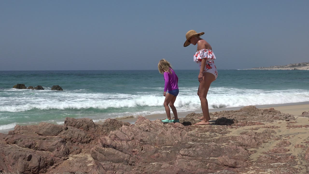 Woman and young girl walking on rocky beach beside ocean surf.