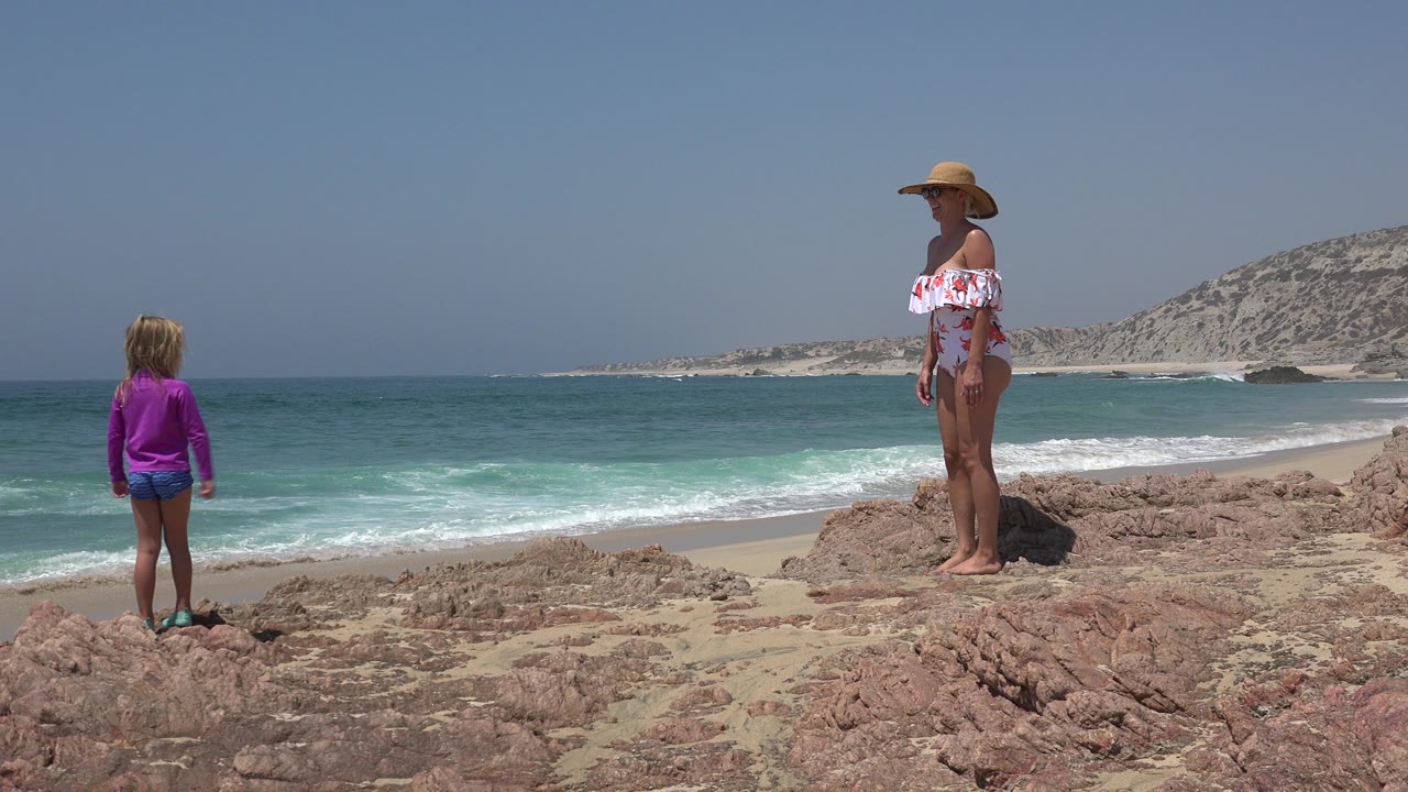 Woman and young girl standing on rocks beside ocean surf.